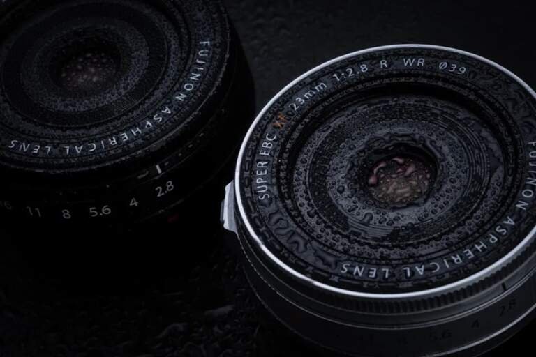 Fujifilm XF 23mm f/2.8 R WR lente pancake compacto con resistencia climática mostrando construcción óptica y marcas de apertura sobre superficie mojada