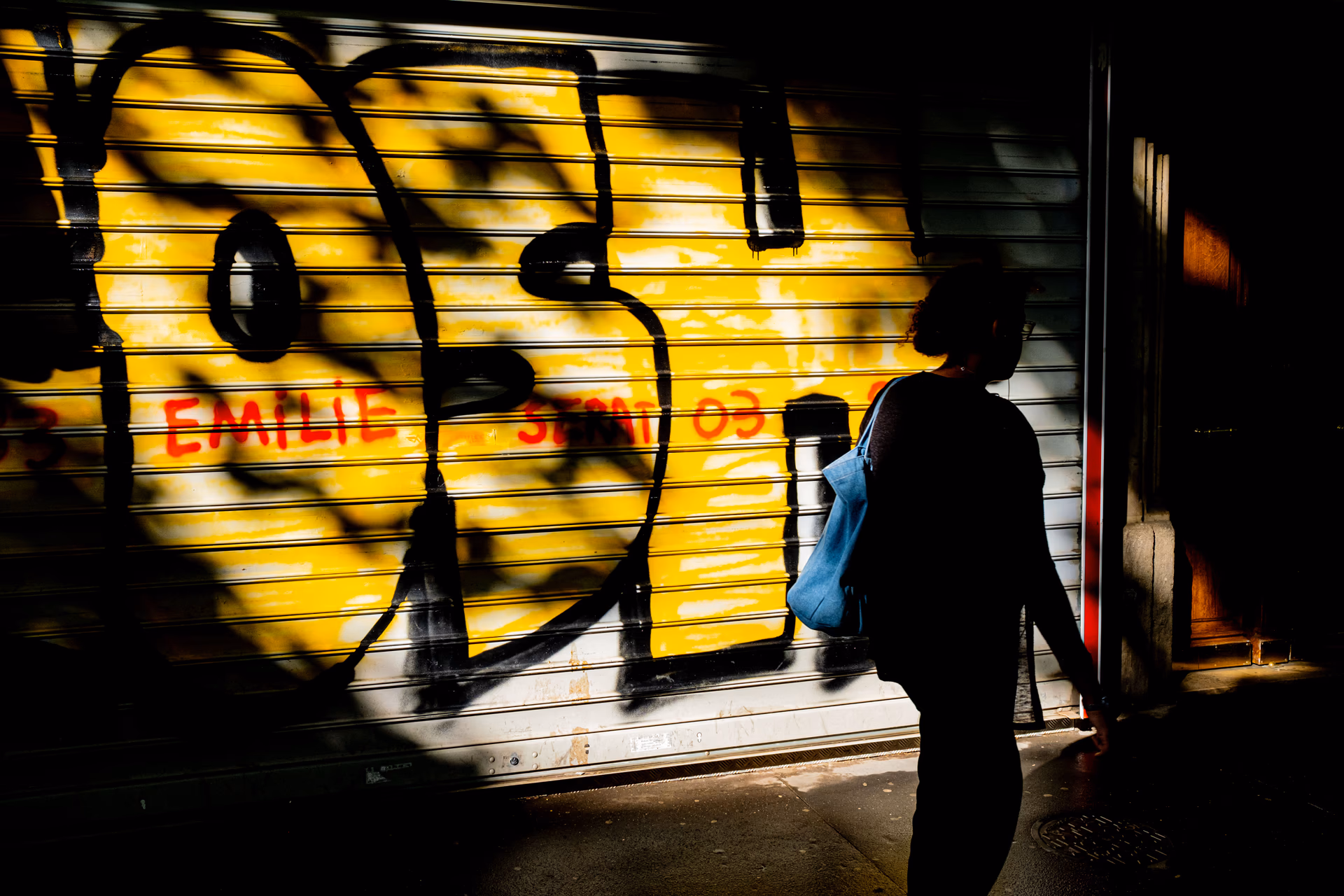 Persona frente a un escaparate iluminado en amarillo con reja metálica y letreros rojos en calle de París — Nikon Zfc TT Artisan 25mm f/2