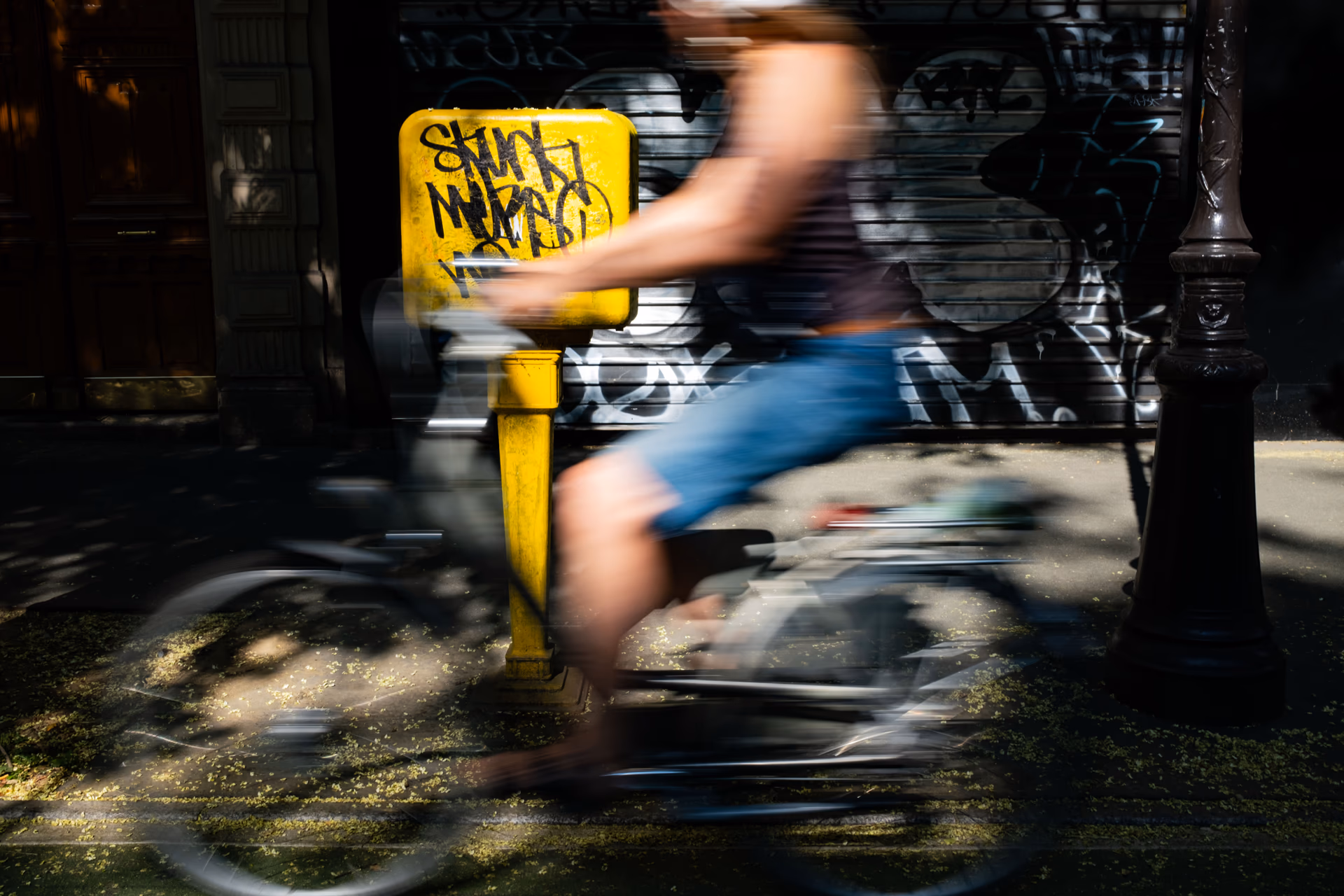 Desenfoque de movimiento: persona en bicicleta pasa junto a un poste amarillo con graffiti en París — Nikon Zfc TT Artisan 25mm f/2