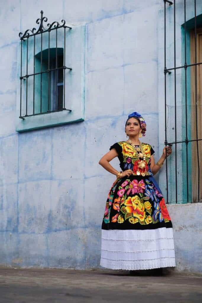 Mujer con vestido tradicional mexicano y flores, frente a pared azul y rejas.