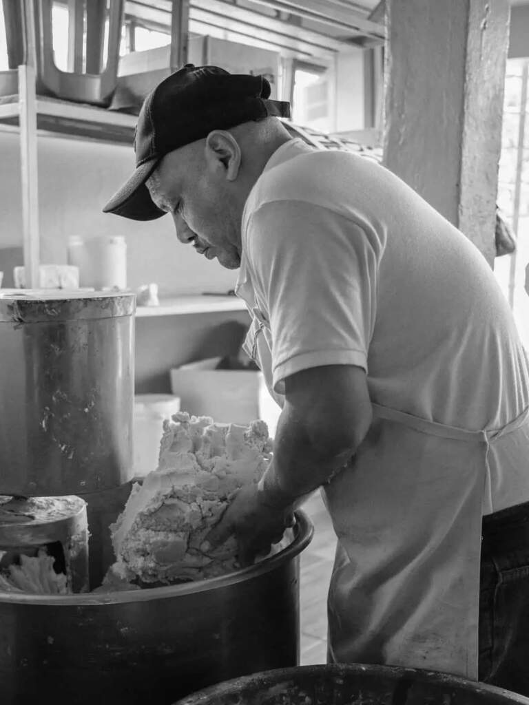 Panadero trabajando masa en panadería mexicana, poca luz y movimiento.