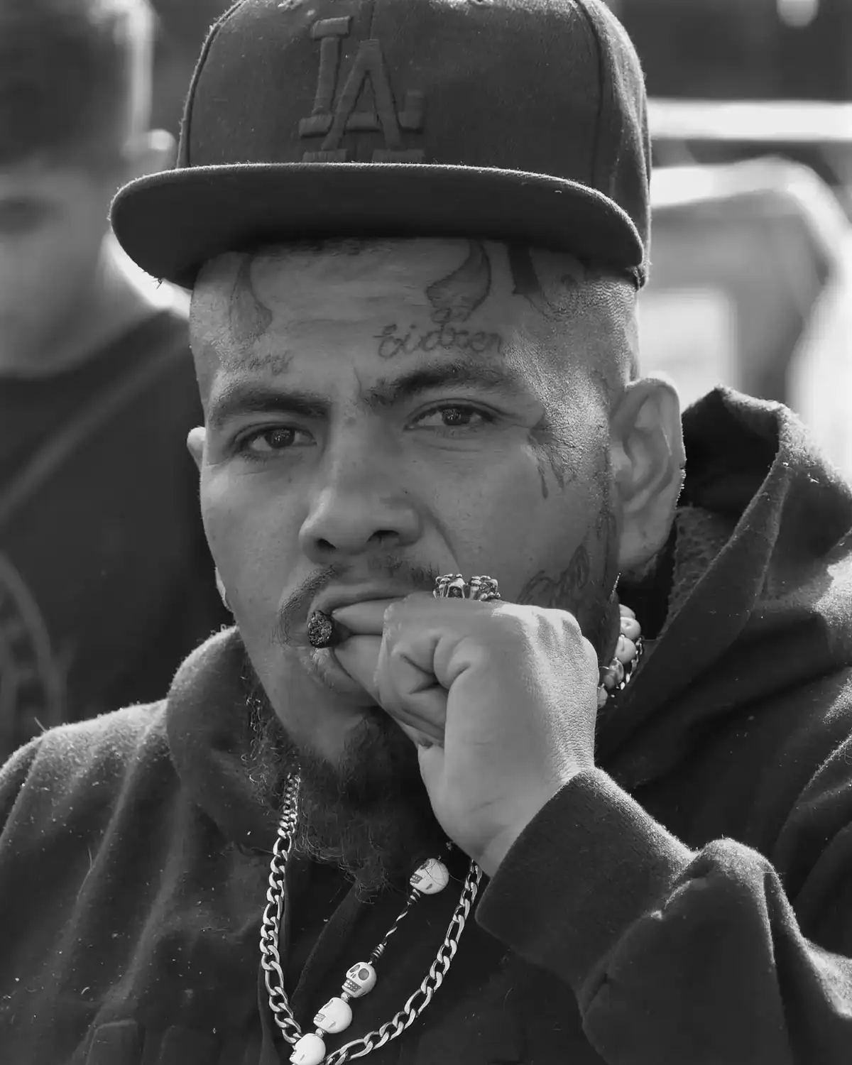 Hombre con tatuajes en la cara y en la frente, que lleva una gorra de Los Ángeles y una cadena con calaveras.