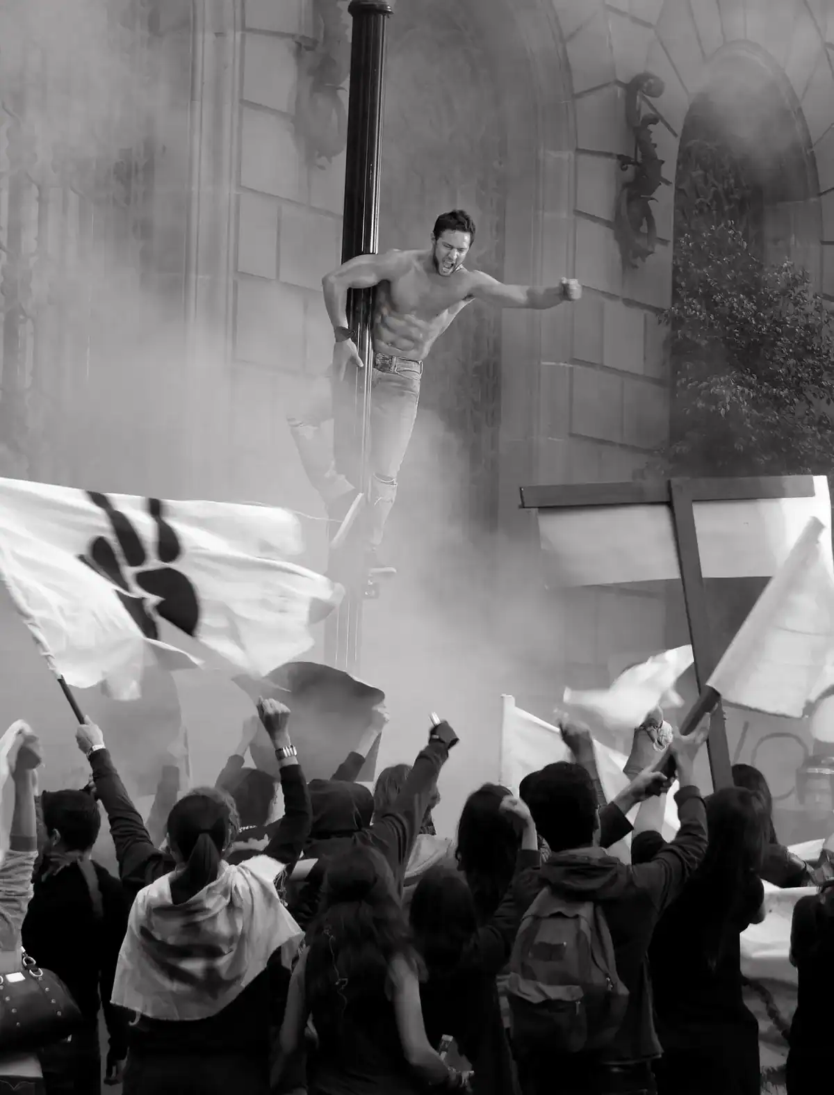 Un hombre con el torso desnudo trepa por una farola, mientras una multitud protesta con banderas.