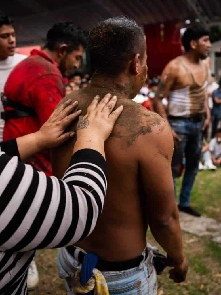 Manos cubren de tierra la espalda de un participante en el palo encebado en Tlaxcala.